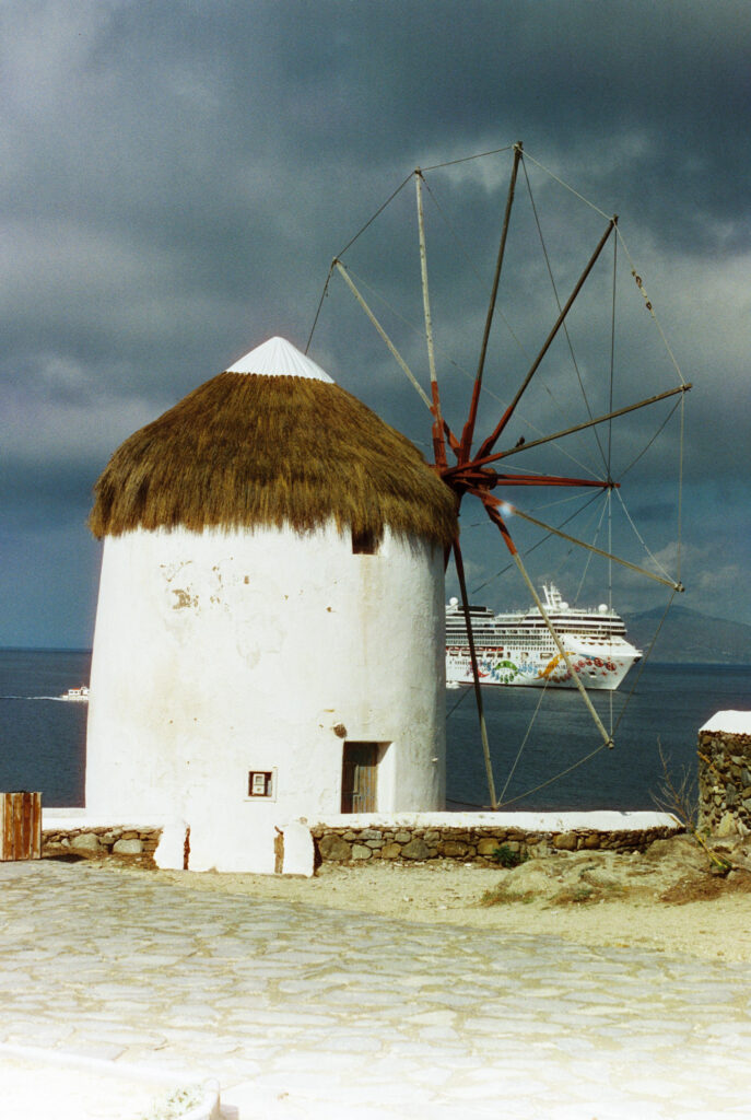 An old windmill in Mykonos with a cruise ship moored in the bay behind it