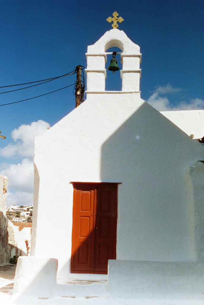 A small whitewashed chapel with an orange wooden door in a Mykonos alley