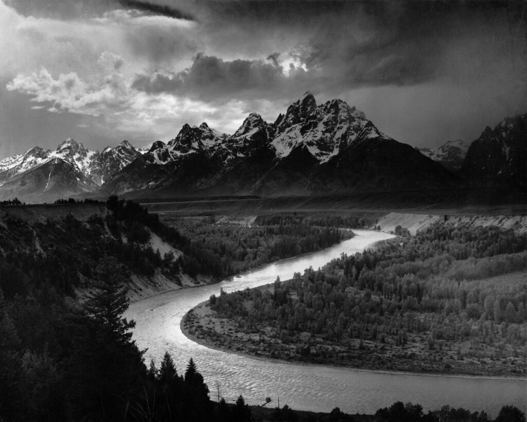 Black-and-white landscape photograph of the Teton mountain range rising in the background above a dark, winding Snake River. The river curves through a broad valley of forests and open land, with dramatic clouds and strong tonal contrast emphasizing the scale and grandeur of the scene.