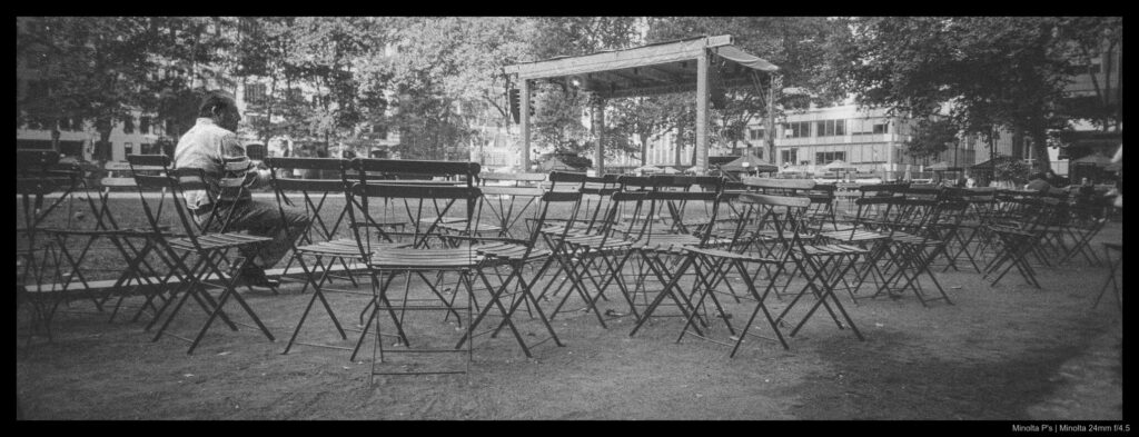 A person sits alone at a metal chair and table in a park, surrounded by other empty metal chairs and tables.