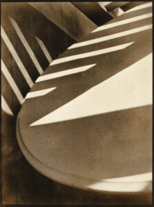 Black-and-white abstract photograph of porch shadows cast across a curved surface and railing. Strong diagonal bands of light and dark form a geometric composition, reducing the scene to interlocking shapes, lines, and tonal contrasts.