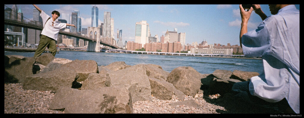 Two friends take a photo in Dumbo, with the Manhattan skyline visible in the background.