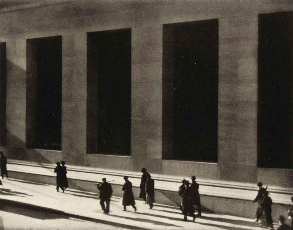 Black-and-white photograph of pedestrians walking past the tall, dark rectangular windows of a Wall Street building. The figures appear small against the massive architecture, creating a stark geometric composition of vertical shadows, bright pavement, and human silhouettes.