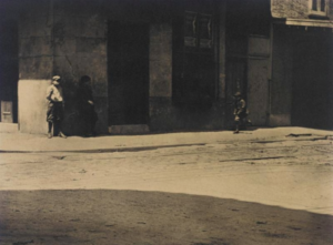 Sepia-toned black-and-white street photograph of a New Orleans building façade in harsh sunlight. Several small figures stand or walk along the sidewalk beneath deep architectural shadows, while a broad empty street and large dark foreground shadow divide the image into stark geometric bands of light and shade.