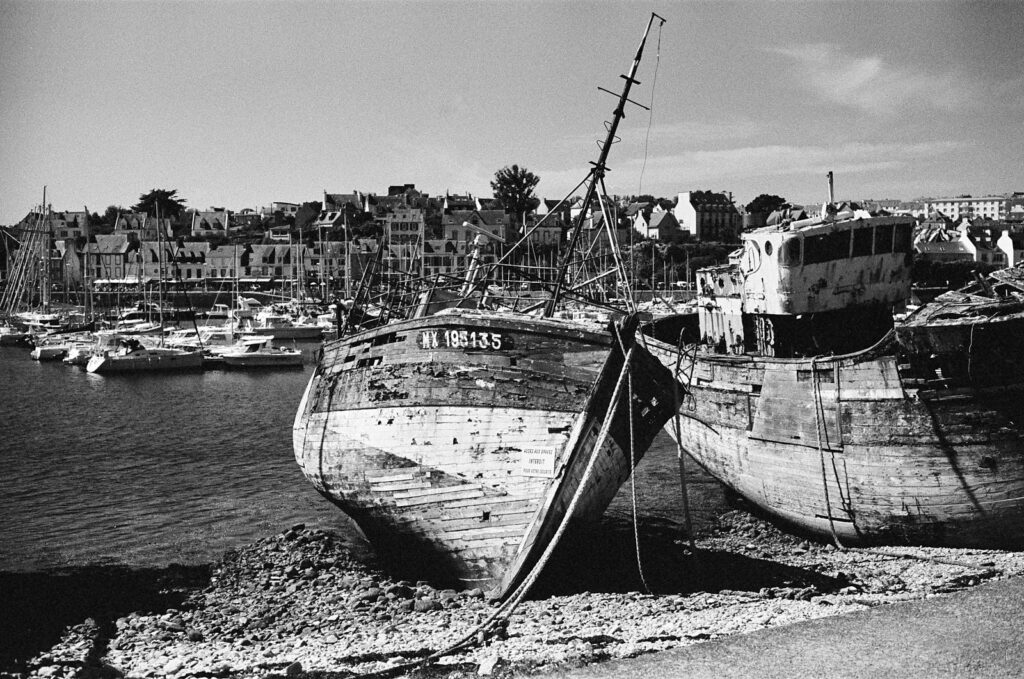 Wider view of a boat cemetery with a Breton town behind, weathered hulls on the shore