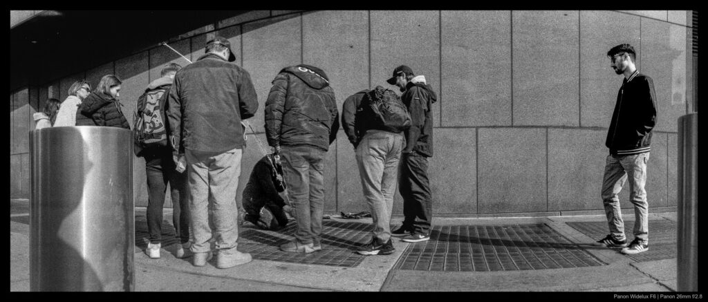A small crowd gathers around a facilities worker using a long flexible rod to attempt to retrieve jewelry that has fallen underneath a grate near the Madison Square Garden building. A new onlooker arrives from the right of the frame.