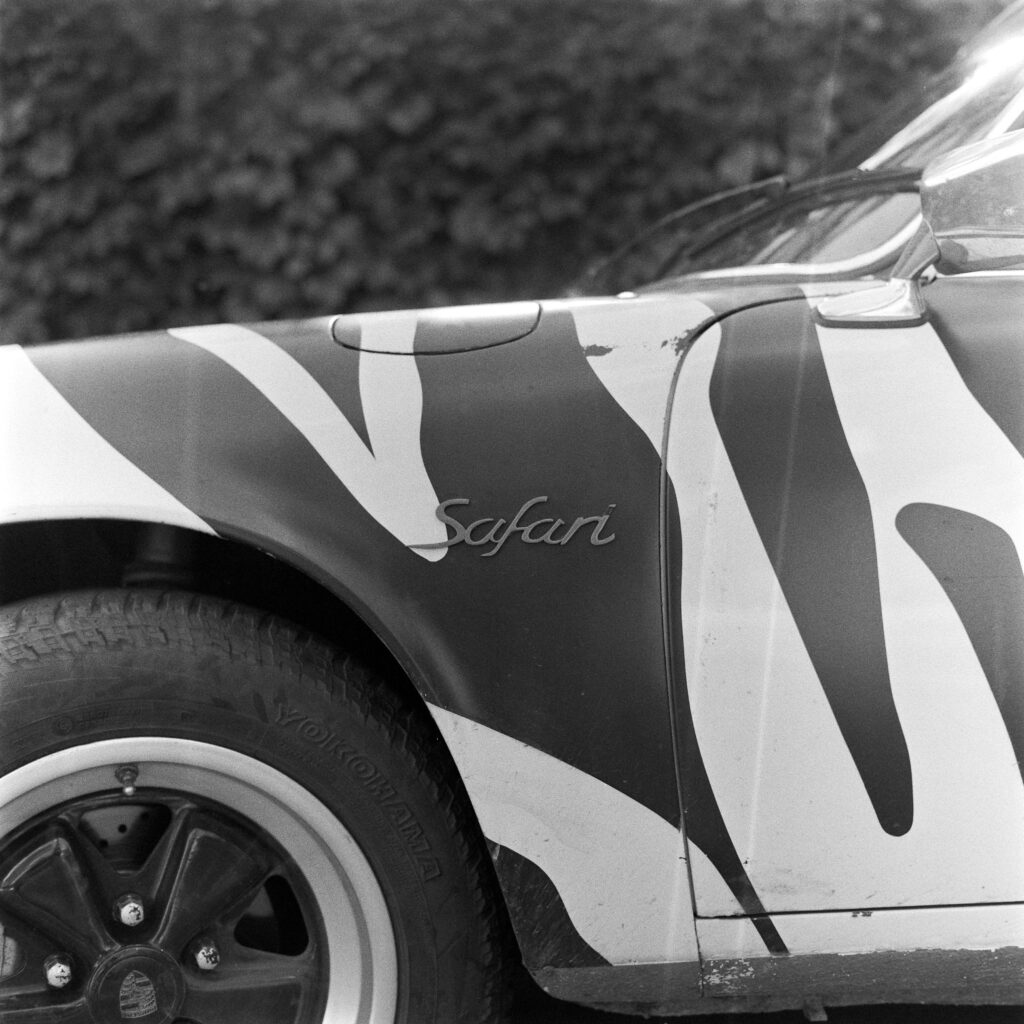 Black and white close-up of the front left fender of a zebra-striped 1974 Safari Porsche 911, featuring a classic script "Safari" badge above a black Fuchs wheel and Yokohama tire.
