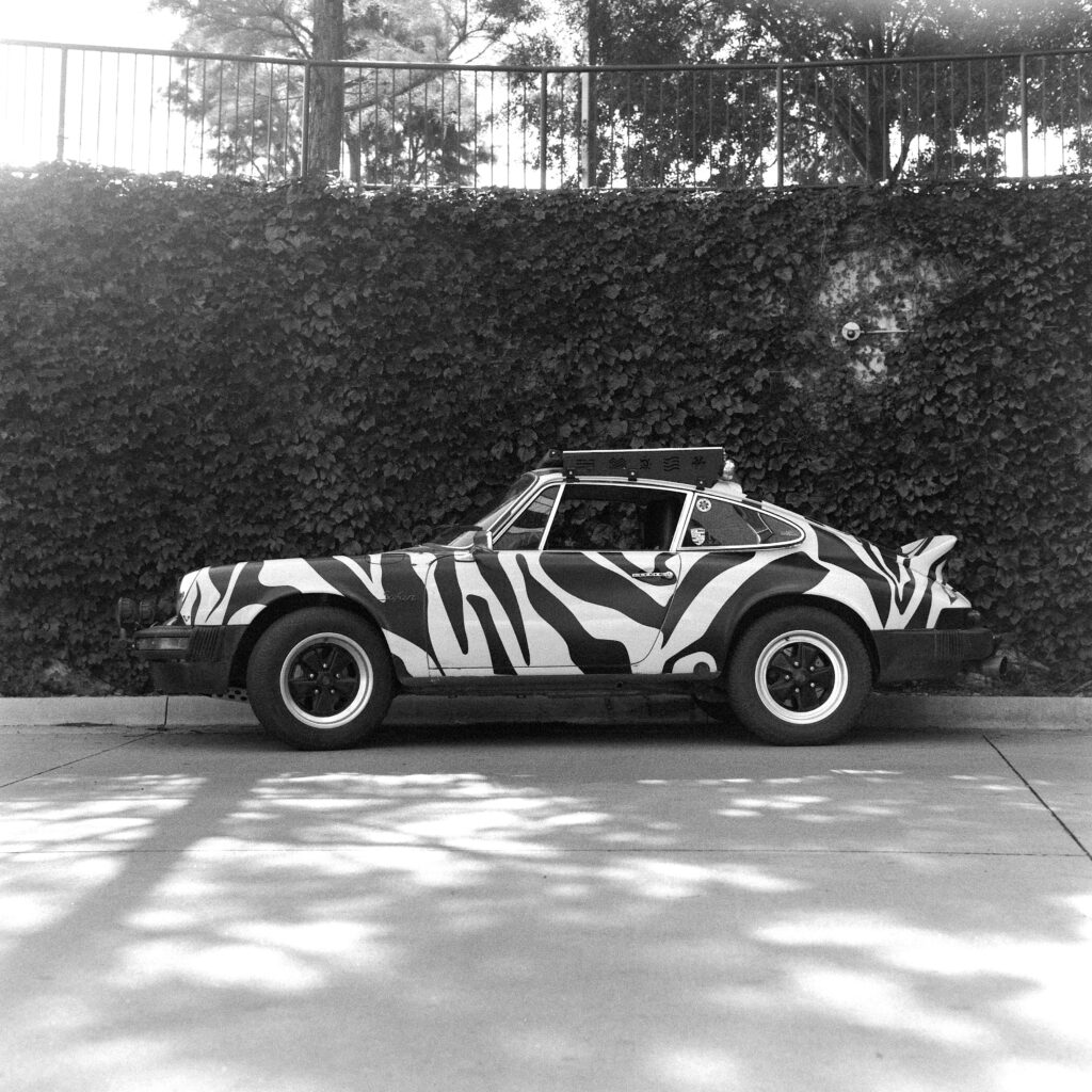Black and white side-profile photo of a lifted 1974 Safari Porsche 911 featuring zebra stripes, a ducktail spoiler, and a roof rack, parked against a wall covered in dense ivy.