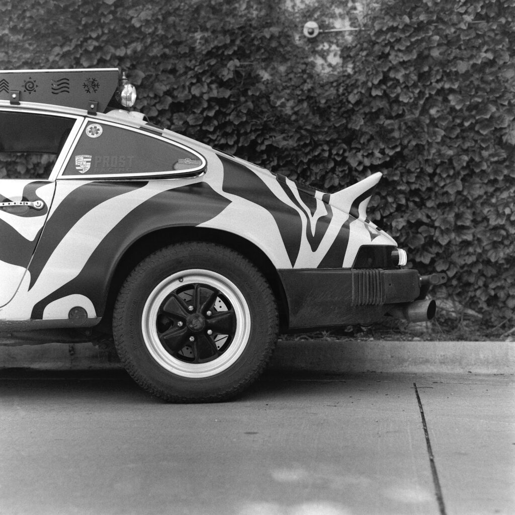 Black and white rear-quarter view of a zebra-striped 1974 Safari Porsche 911 showing the classic ducktail spoiler, rear wheel, and custom roof rack against an ivy wall.