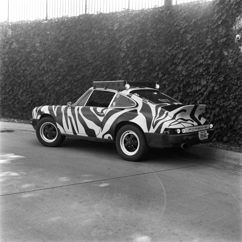 Black and white angled rear view of a lifted 1974 Safari Porsche 911 with a zebra wrap, roof rack, ducktail spoiler, and a Kansas Antique license plate.