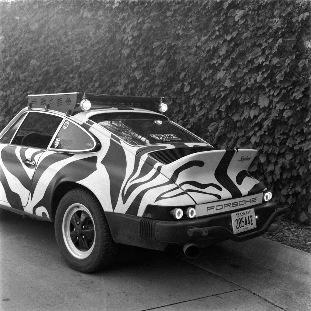 Black and white close-up of the rear end of a 1974 Safari Porsche 911, highlighting the Kansas Antique license plate, rear bumper lights, ducktail spoiler, and a roof rack with engraved symbols.