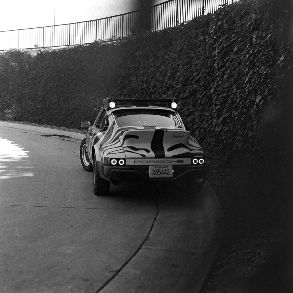 Black and white photograph of a zebra-striped 1974 Safari Porsche 911 viewed from the rear, parked on a curved driveway next to a tall ivy-covered wall, with a soft out-of-focus element in the right foreground.