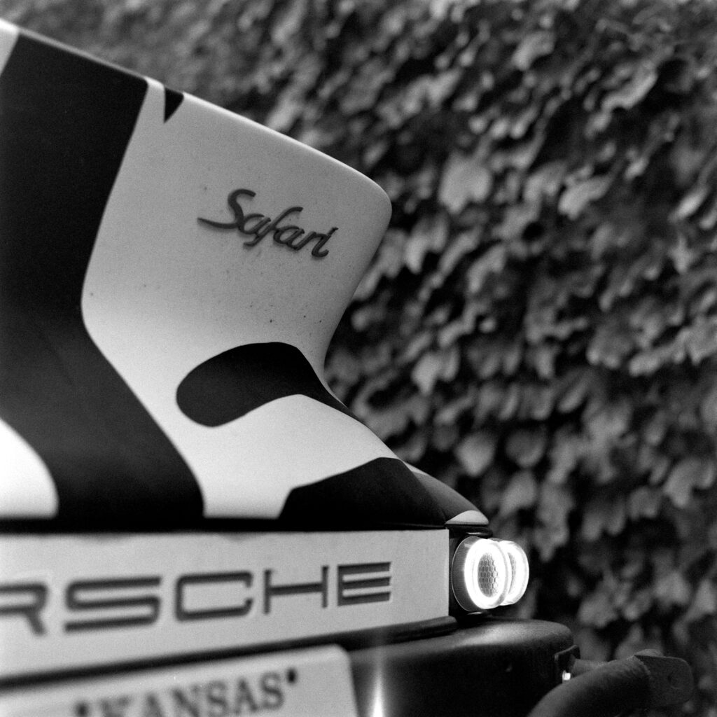 Black and white close-up of the rear ducktail spoiler of a 1974 Safari Porsche 911 featuring a "Safari" script badge, zebra livery, and glowing circular taillights above a Kansas license plate.