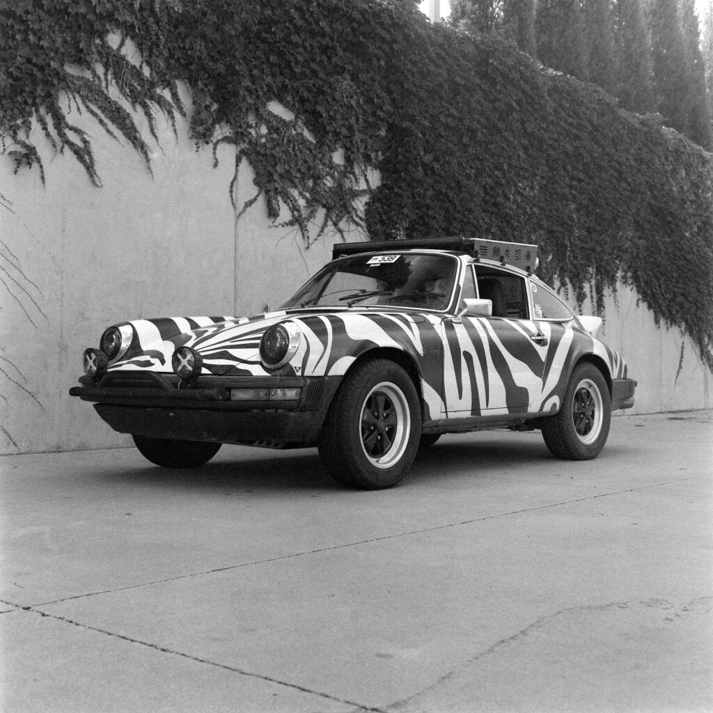 Black and white three-quarter view of a lifted, zebra-striped 1974 Safari Porsche 911 parked on concrete in front of a smooth wall draped with creeping ivy.