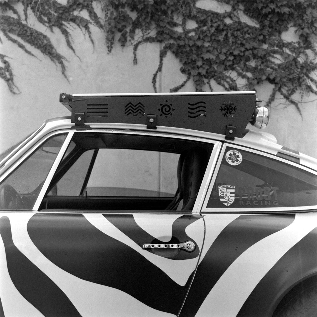 Black and white close-up of the roofline of a zebra-striped 1974 Safari Porsche 911, showing a black custom roof rack with cutout elemental symbols and window decals below.