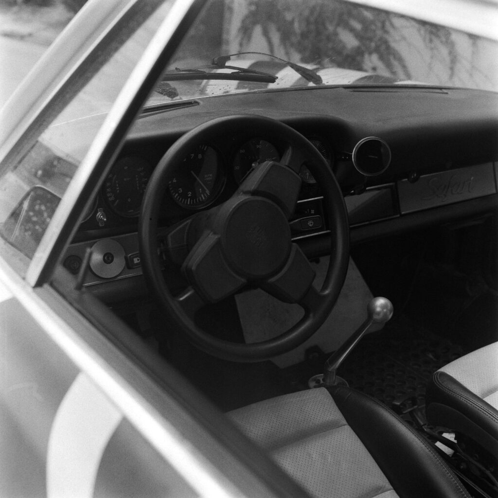 Black and white interior shot of a 1974 Safari Porsche 911 viewed through the driver's window, showing the steering wheel, classic analog gauges, tall manual shifter, and a "Safari" badge on the passenger dash.