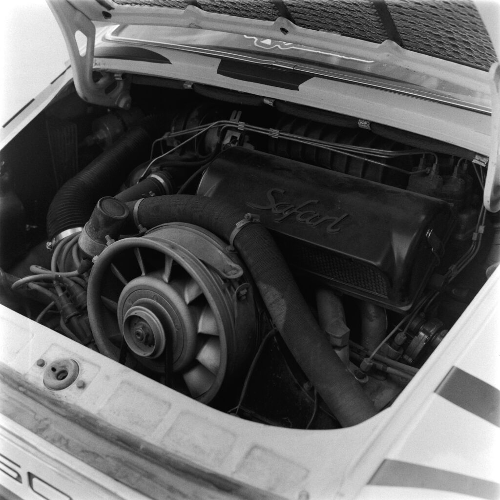 Black and white overhead close-up of the open rear engine bay of a 1974 Safari Porsche 911, displaying the flat-six engine and a custom black cover embossed with "Safari".