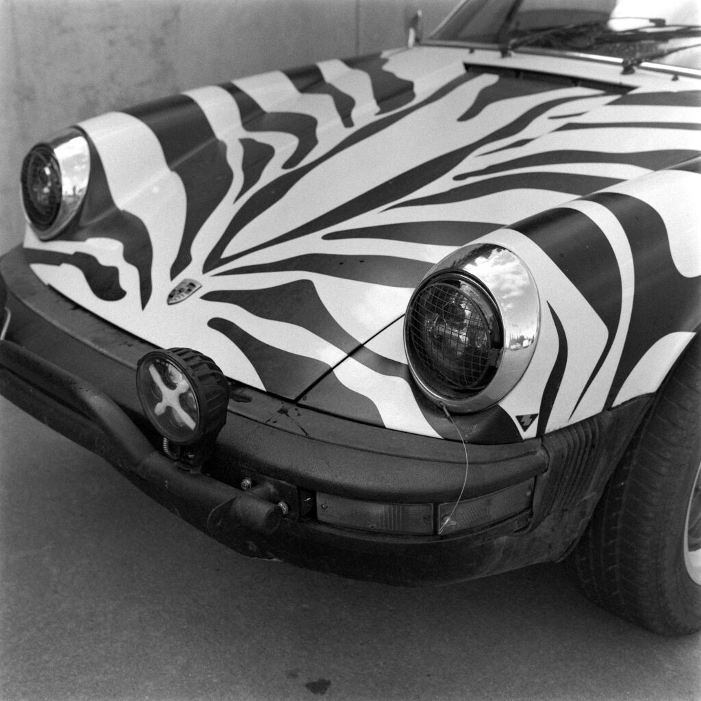 Black and white close-up of the front passenger-side hood and fender of a zebra-striped 1974 Safari Porsche 911, showing a mesh-covered headlight and an auxiliary light mounted to a tubular bumper.