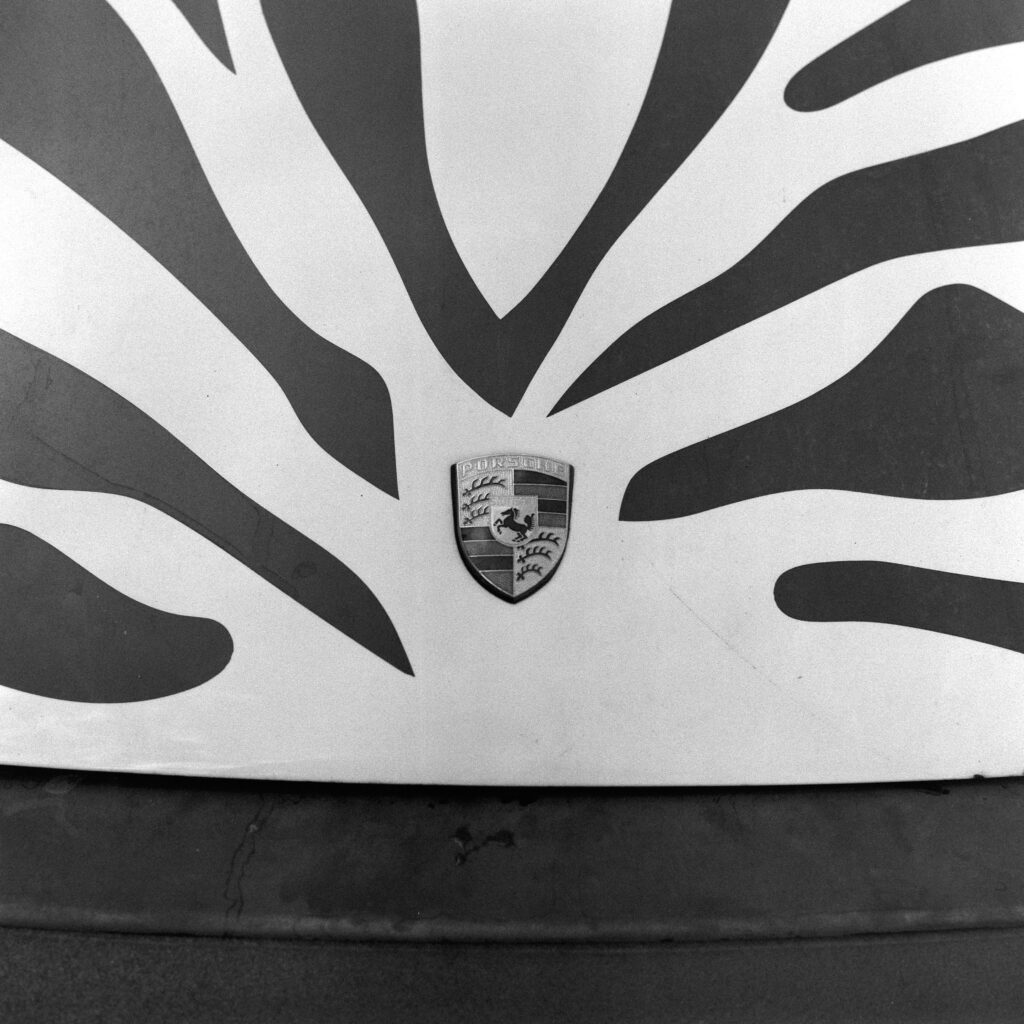 Black and white overhead close-up of the front hood of a 1974 Safari Porsche 911, showing black zebra stripes converging symmetrically toward the central Porsche emblem.