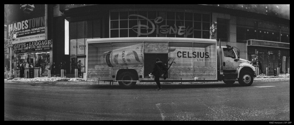 A truck crewman lifts a dolly out of a large delivery truck's side hatch in preparation to unload cargo. The Disney flagship store logo is visible looming over the truck, which is branded with the name "Celsius" and displays a promotional image of a beverage can.