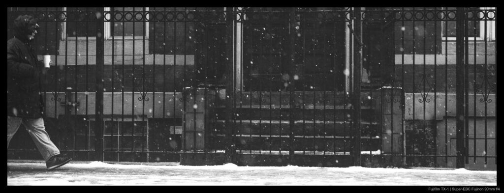 A person holding a coffee walks by a fenced-off residence during a moderate snowfall.