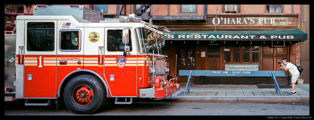 A tourist bends forward with a camera in their hands, pointed at the front of a large FDNY fire truck parked a short distance away. A wooden police barrier is situated lengthwise between the two and a restaurant is visible in the background.