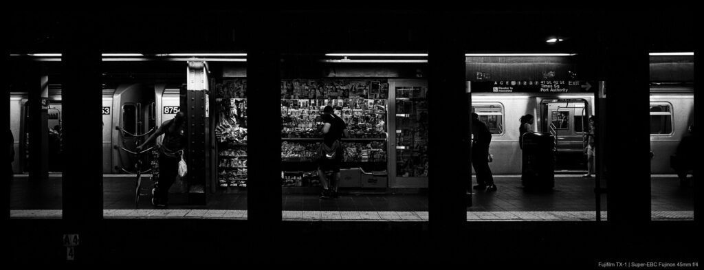 Support columns in a subway station divide a series of small scenes of people standing around a station vending kiosk, benches, and the platform in general in a way that evokes a strip of exposed film.