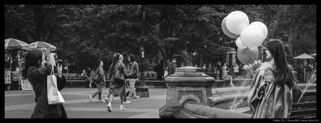 A person captures their friend's graduation day cap and gown with a camera, aiming across the frame towards the fountain at Washington Square Park.