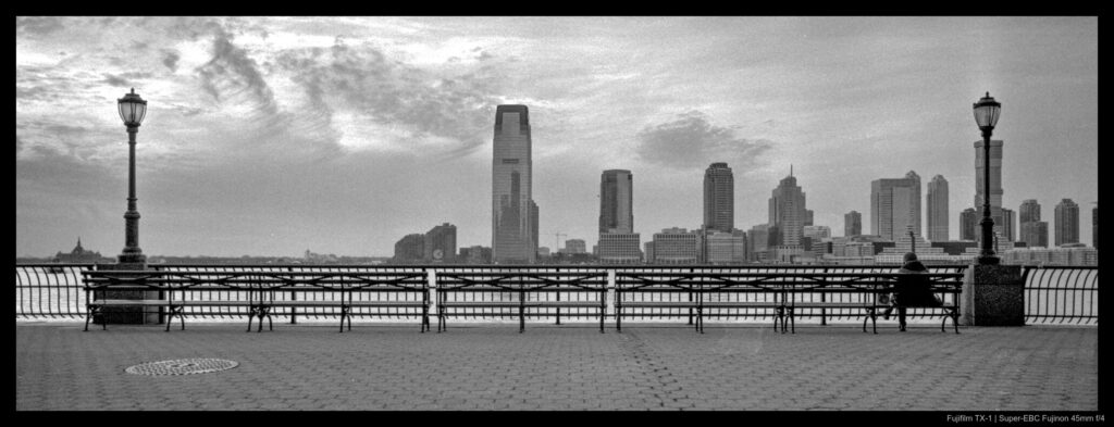 A person sits on the right-hand edge of a long row of benches, situated between two tall lampposts. The scene overlooks the Hudson River, with the New Jersey skyline visible in the background beneath a cloudy sky.