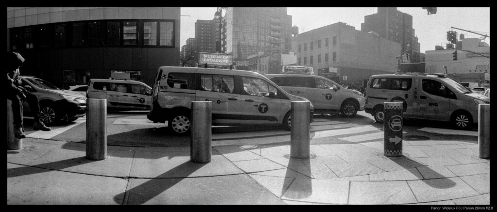 A person sits on a bollard facing to the right next to a one-way street filled with taxis driving to the right.
