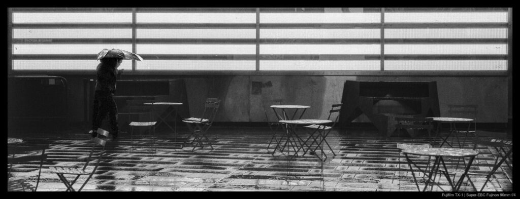 A person beneath an umbrella walks by a large installation of LEDs in Times Square, their glow silhouetting them and reflecting off of the wet concrete ground. Empty tables and chairs are scattered about the area.