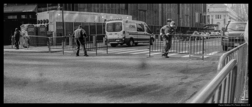 Two workers slide metal barricades aside to open a roadway near Madison Square Garden. The two are moving in opposite directions, with one facing towards the camera and the other turned away.