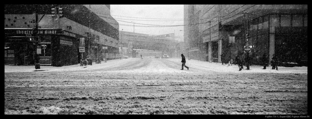 A person crosses a street covered in churned-up snow as more continues to fall from the sky. One block further down, another person is visible making the same crossing, their legs in perfect sync with the figure in the foreground.