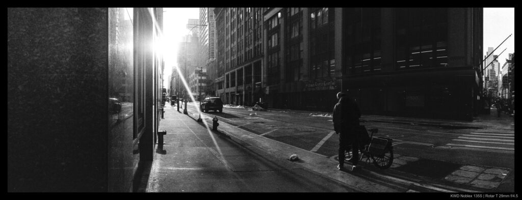 A bicyclist prepares to mount their bicycle beneath a dramatic morning light cast down the length of a NYC street.