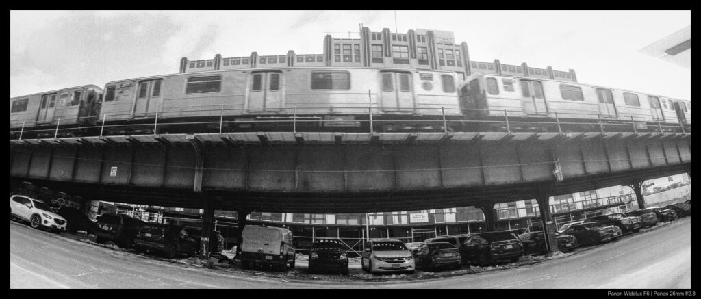 A subway makes its way down an elevated rail line, beneath which is parked a multitude of cars that stretch from left to right beneath the train. Mid-rise buildings are visible in the background.