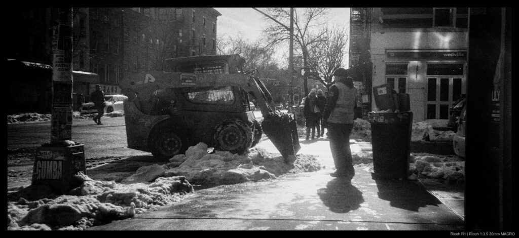 A worker supervises a small vehicle scraping compacted snow away from a curbside.