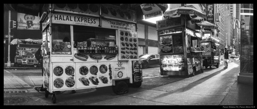A prospective customer looks at one food cart in a line of three located in Times Square, casting a long shadow that stretches from the rear cart to the one in the foreground.