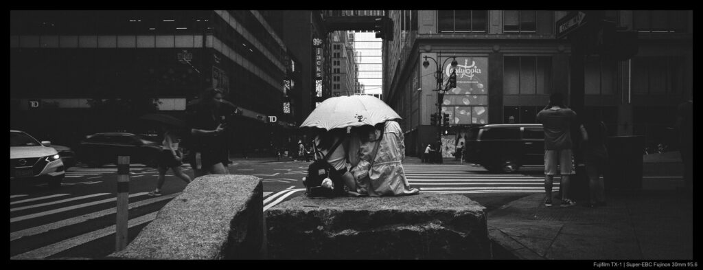 Two people shelter beneath an umbrella during a rainy moment in Herald Square, seated on a large stone block. Other buildings are visible in the background, across the street towering up and out of sight.