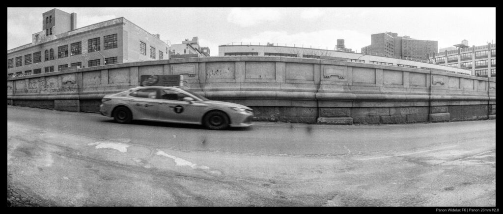 An image of a taxi driving in front of a ramp, with midrise buildings visible in the background. The car is slightly elongated due to the motion of the camera lens.
