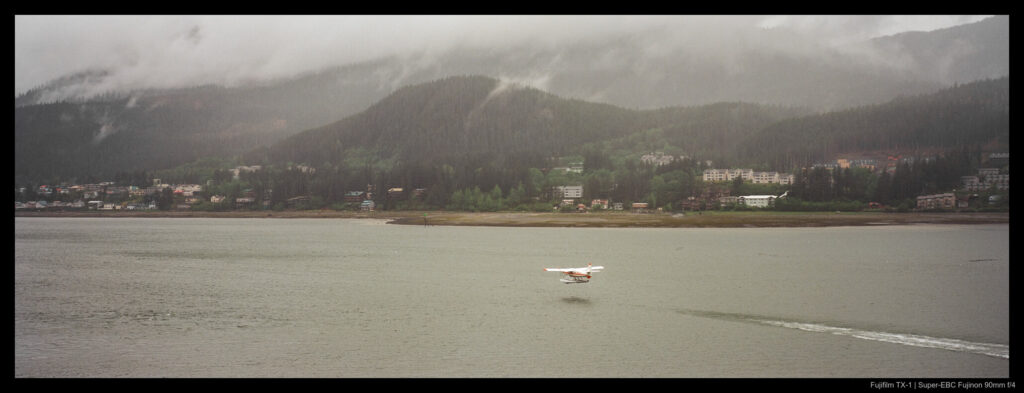 A seaplane taking off from an inlet in Alaska. Fog and clouds shroud mountains across the water, as well as buildings interspersed with thick growths of trees. A wake left by the airplane is visible trailing the craft.