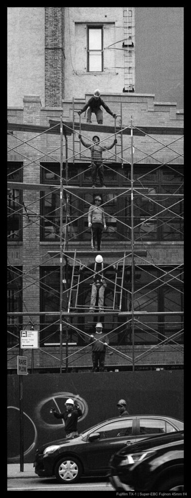 Five workers stand in a vertical line on different levels of scaffolding, passing materials up the line. Another worker is visible on the other side of a fence on the ground.