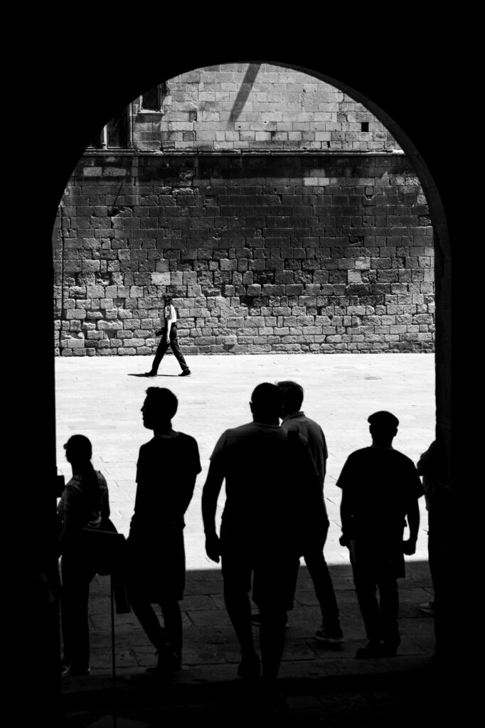 Silhouetted figures under a stone archway in Barcelona's Gothic Quarter, with a lone figure crossing a sunlit square beyond