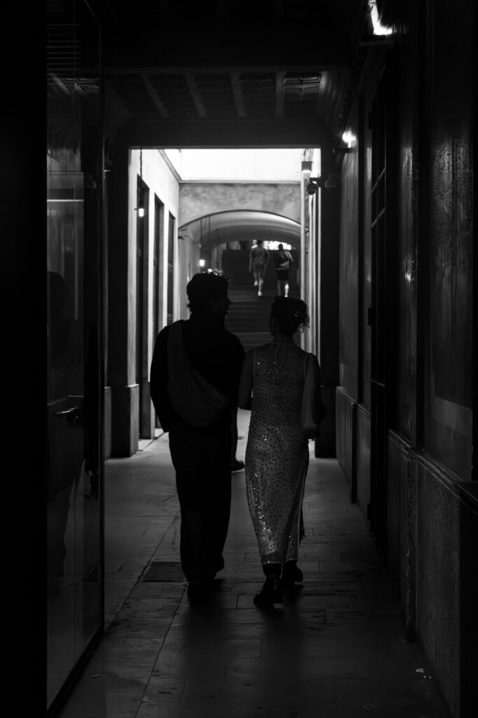 A couple walking arm in arm through a dimly lit arcaded passageway at night, two more figures visible ahead