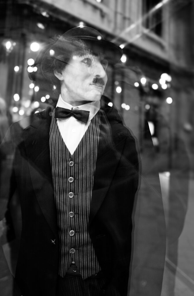 A shop-window mannequin dressed as Charlie Chaplin, reflected among string lights and passersby at night