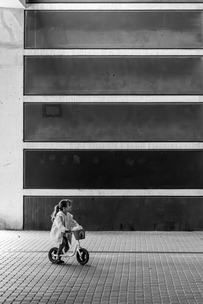 A young girl riding a balance bike past a dark, reflective panelled building facade