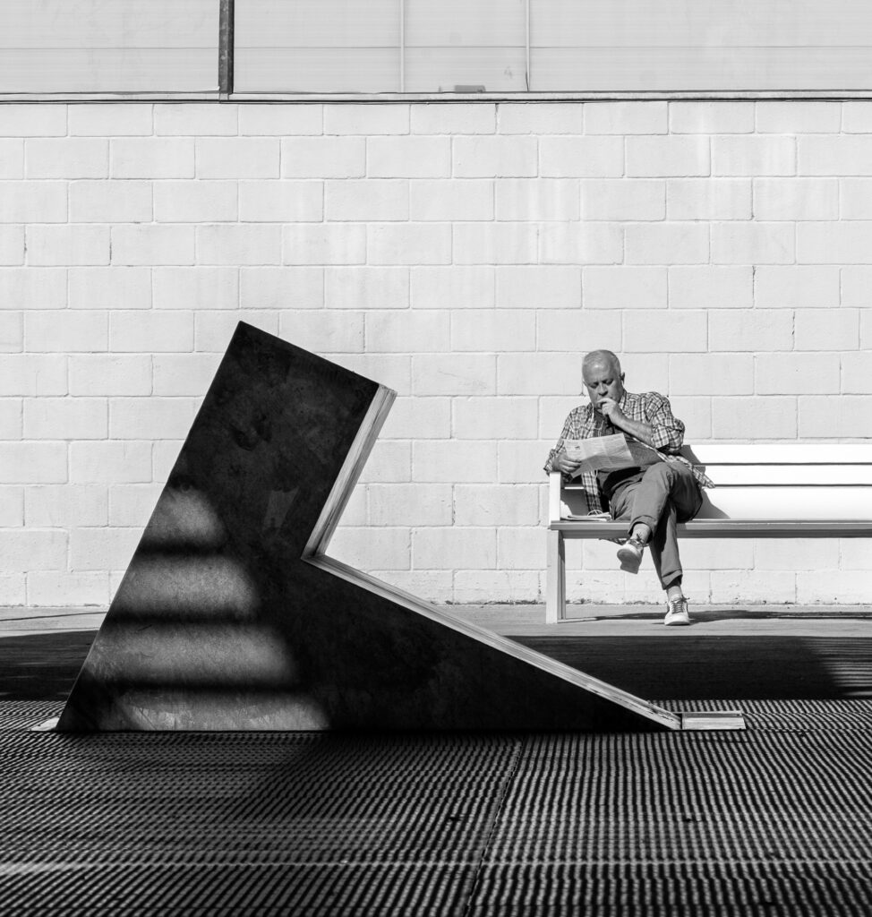 A man reading on a bench beside an angular black triangular sculpture against a white brick wall