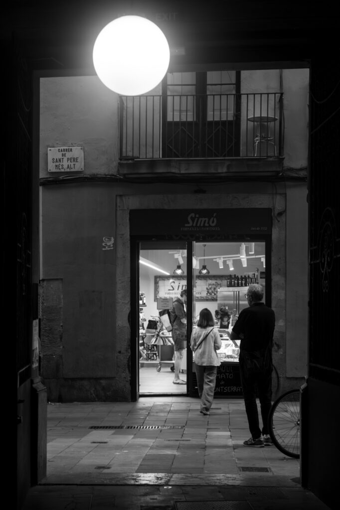 A glowing round lamp above the entrance to a lit cheese shop at night, with customers inside