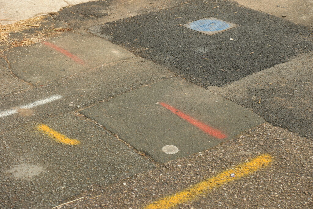 A photograph showing a pavement with drying patches and coloured marks in red, blue and yellow paint.