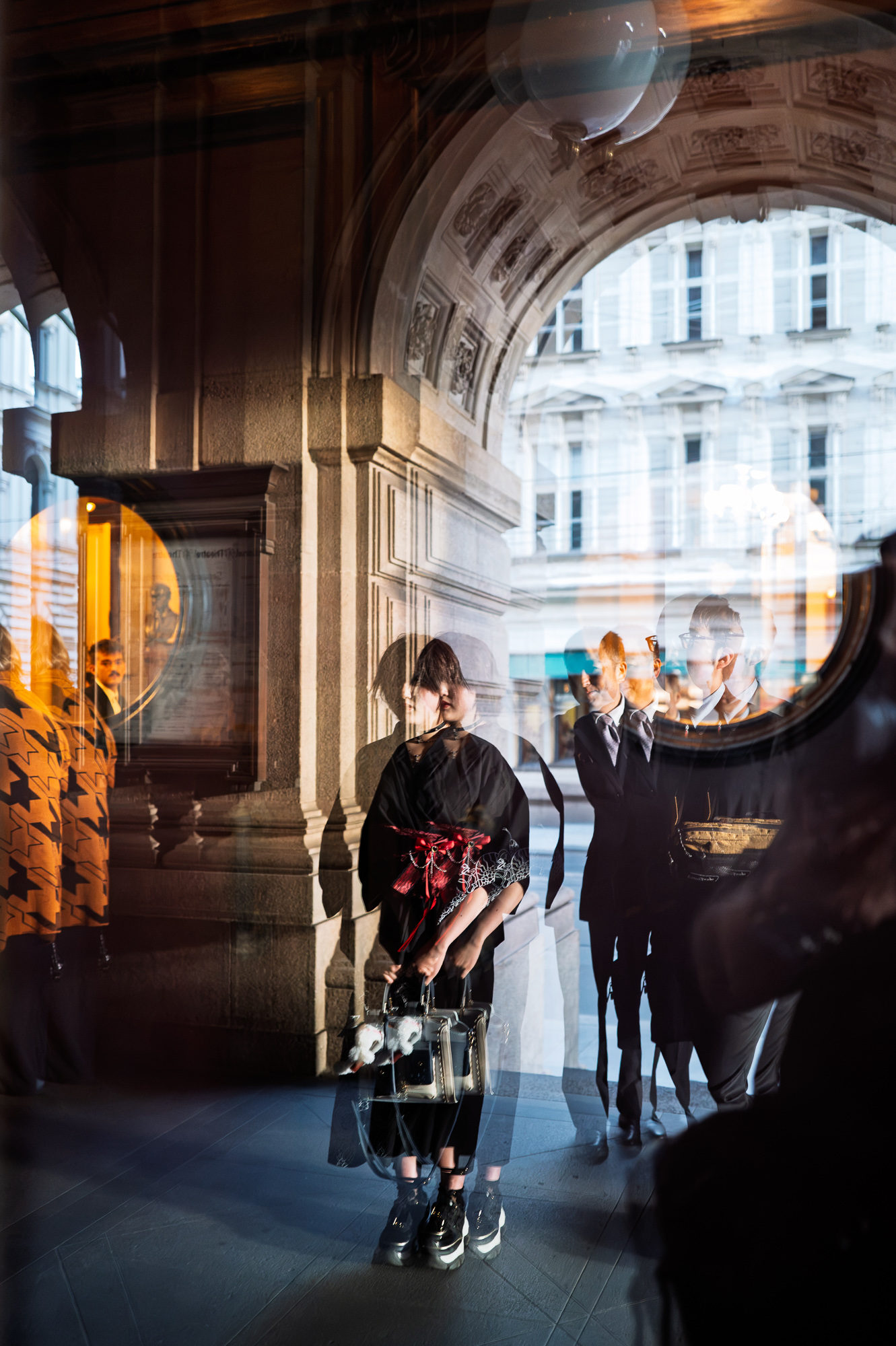 Young woman in black with a red bow reflected in the glass doors of Prague's National Theatre, among layered arches and façades.